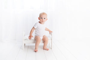 A toddler sits upright on a small white bed, wearing a white tshirt and nappy, looking straight into the camera with big blue eyes.. The floor and walls are white.